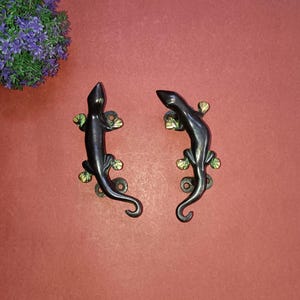 Op de afbeelding: Twee zwarte metalen deurgrepen in de vorm van een hagedis met groene accenten. De hagedissen kijken in tegengestelde richtingen.