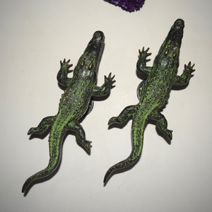 May include: Two green plastic toy crocodiles with black eyes and gold accents. The crocodiles are posed with their mouths open and their tails curled.