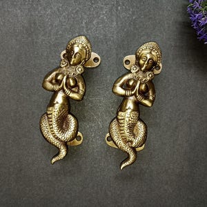 May include: Two antique brass mermaid door pulls. Each mermaid figure has a detailed design, with hands clasped in prayer and a scaled tail. The door pulls are mounted on a dark gray surface, with a small cluster of purple flowers in the background.