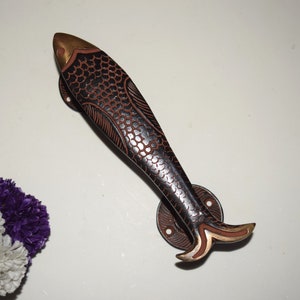 May include: A black and brown fish-shaped door handle with a gold accent on the tail. The handle is mounted on a round, black metal plate with three screws.