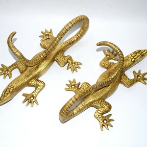 Pode incluir: Dois ganchos de metal de cor dourada em forma de lagarto. Os lagartos estão enroladas com suas caudas formando um gancho. Os ganchos são decorativos e seriam ótimos para pendurar chaves, toalhas ou outros itens.