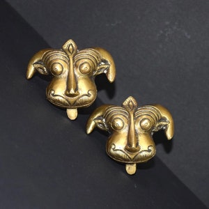 May include: Two gold-coloured metal decorative ornaments shaped like animal faces with intricate details. The ornaments have a textured surface and a slightly raised design.