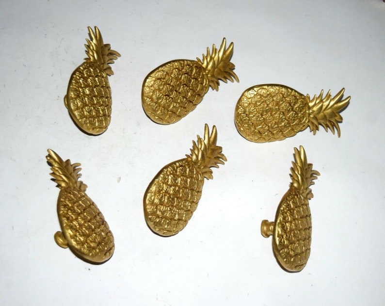 May include: Six gold pineapple-shaped drawer pulls. The pulls are made of metal and have a shiny finish.