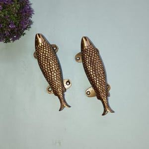May include: Two bronze-coloured fish-shaped door handles. Each handle has a textured scale pattern and is mounted with two small screws. The handles are designed to be mounted horizontally. Decorative hardware.