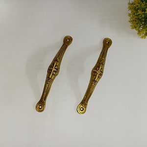May include: Two gold-toned metal door handles with a decorative face design. The handles are long and narrow with a rounded end.