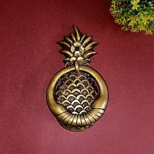 May include: A brass pineapple door knocker with a ring handle. The knocker features a detailed design of a pineapple, with a textured surface and a ring for knocking. The door knocker is a decorative item.