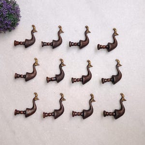 Può includere: Una collezione di dodici ganci decorativi a forma di pavone. I ganci sono color bronzo con accenti dorati sulla testa e sulla coda. Sono disposti in tre file da quattro su una superficie bianca.