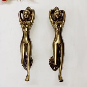 Puede incluir: Un par de manijas de puerta de latón a juego, cada una con la figura esculpida de una persona con los brazos levantados sobre la cabeza. El acabado metálico tiene un tono dorado antiguo cálido con detalles oscuros y cada manija incluye una base.