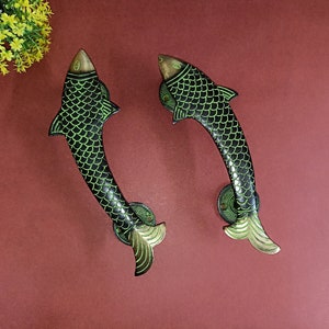Op de afbeelding: Twee decoratieve deurgrepen in de vorm van vissen. De handgrepen zijn zwart met groene accenten, waaronder de schubben, vinnen en ogen. De visvormige handgrepen zijn gemaakt van metaal en staan tegen een donkerrode achtergrond.