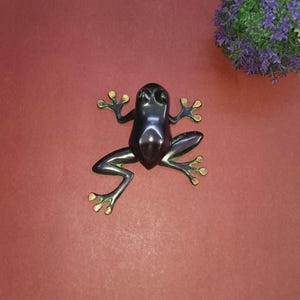 Könnte beinhalten: Schwarze Froschfigur mit grünen und goldenen Akzenten. Der Frosch ist mit ausgestreckten Beinen und leicht nach oben geneigtem Kopf posiert.