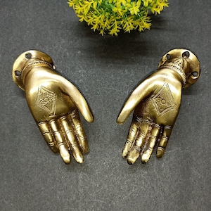May include: A pair of metallic gold-toned wall hooks shaped like open hands. Each hand features a circular mounting base with two screw holes and an engraved diamond-shaped floral pattern centered on the palm with detailed finger articulation.