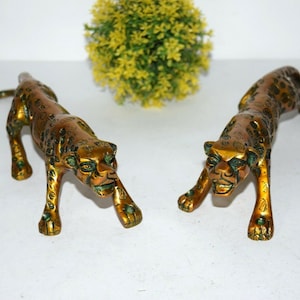 May include: Two gold-colored metal leopard figurines with green spots. The leopards are in a crouching position with their heads facing forward.