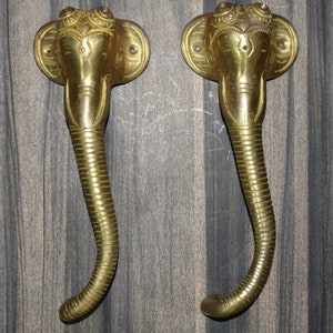 May include: A pair of gold-toned door handles shaped like elephant heads with intricate details. The handles are mounted on a dark wood surface.