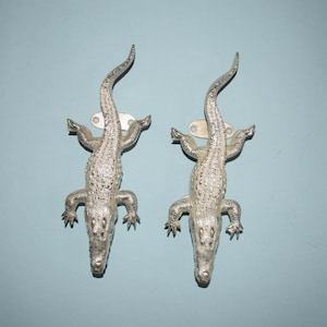 May include: Two silver alligator-shaped wall hooks. The hooks are mounted on a pale blue wall.