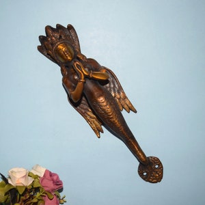 May include: A gold-toned metal door handle shaped like a mermaid with wings. The mermaid is holding her hands together in a prayer position. The handle has a detailed design and is a unique and decorative piece.