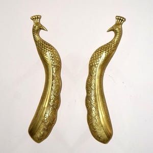 Może przedstawiać: Dwa mosiężne uchwyty do drzwi w kształcie pawia. Uchwyty są w kolorze złotym i mają skomplikowane detale, w tym teksturowany korpus i koronopodobny wzór na głowie. Uchwyty są zakrzywione i przeznaczone do chwytania.