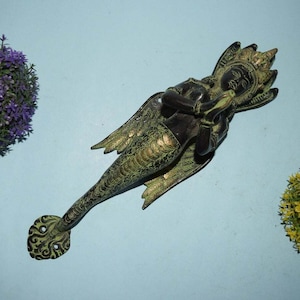 Puede incluir: Un llamador de puerta de metal verde y dorado con forma de sirena con alas. La sirena tiene las manos juntas en posición de oración.