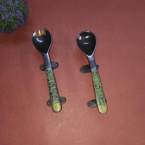 Puede incluir: Un juego de dos utensilios para servir: un tenedor y una cuchara. Los utensilios son negros con detalles decorativos verdes y dorados en los mangos. El tenedor tiene cuatro púas. La cuchara tiene un cuenco redondeado.