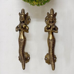 以下が含まれることがあります： 祈りのポーズで手を合わせた女性の形をした、真鍮色のドアハンドルが2つ。ハンドルは、頭飾りや鱗状の下半身など、詳細なデザインが施されています。白い表面に取り付けられています。
