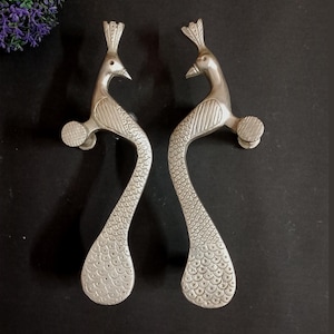 Può includere: Due maniglie per porte argentate a forma di pavone. Le maniglie presentano dettagliati motivi a piume e un pomello circolare all'estremità. Le teste dei pavoni hanno una cresta dettagliata. Progettate per un uso decorativo e funzionale.