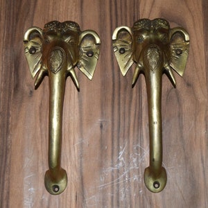 Könnte beinhalten: Zwei verzierte Messing-Türgriffe in Form von Elefantenköpfen. Die Griffe sind mit aufwendigen Schnitzereien verziert und haben eine polierte goldene Oberfläche.