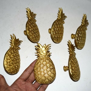 May include: Six gold pineapple-shaped cabinet knobs. The knobs have a detailed pineapple design and are made of metal.