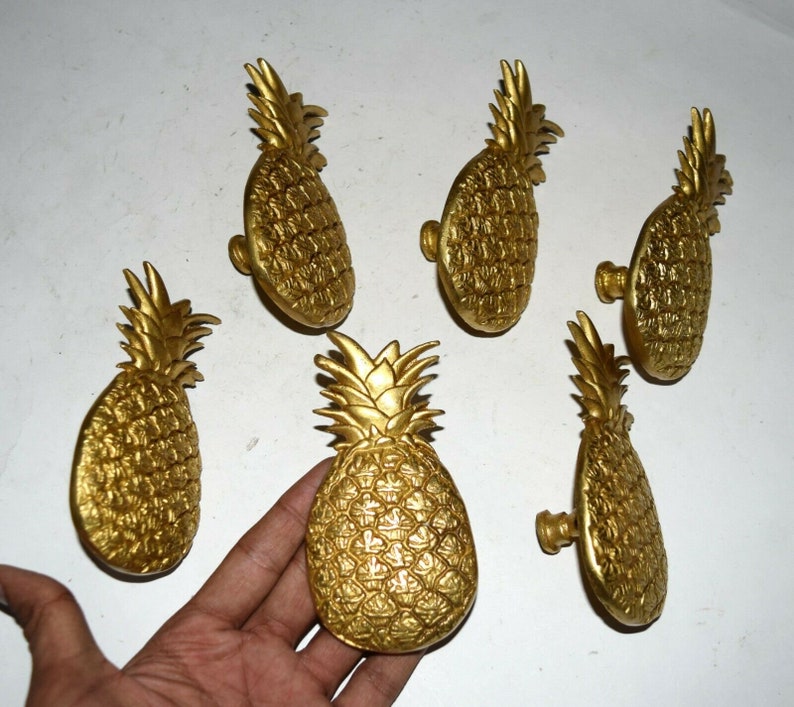 May include: Six gold pineapple-shaped cabinet knobs. The knobs have a detailed pineapple design and are made of metal.