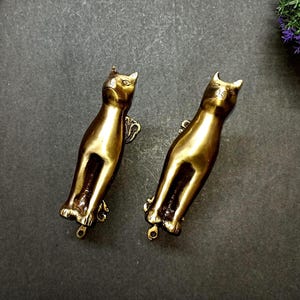 以下が含まれることがあります： 真鍮製の猫の形をしたアンティークドアノッカー2つ。ノッカーは、尖った耳と取り付け用の小さなループを備えた、洗練された細長いデザインで詳細に表現されています。真鍮は、ヴィンテージの職人技を示唆する、暖かく、古くなった緑青を持っています。