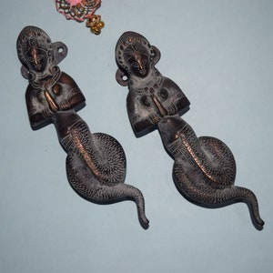 May include: Two ornate bronze door handles shaped like women with snake tails. The women are wearing elaborate jewelry and have their hands clasped in prayer.