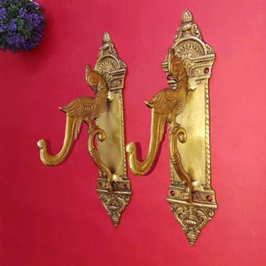 May include: Two matching brass wall hooks featuring an ornate vertical backplate with intricate engravings and a protruding bird-shaped hook. Each piece displays a polished metallic gold finish with detailed carvings at the top and bottom ends.