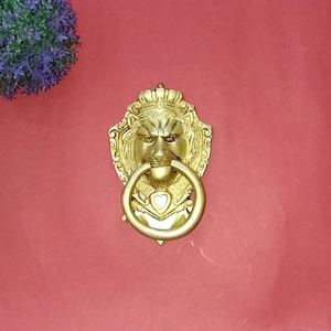 Peut inclure: Un heurtoir de porte en forme de tête de lion doré avec une poignée en anneau. Le lion porte une couronne et un détail en forme de cœur sous l'anneau. Le heurtoir est monté sur une surface rouge, avec une plante violette en haut à gauche.