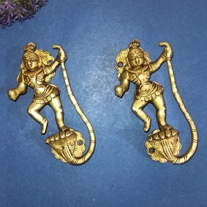 Op de afbeelding: Twee gouden metalen decoratieve wandhaken in de vorm van een dansend figuur dat een gebogen staaf vasthoudt. De figuren staan op een lotusbloembasis.