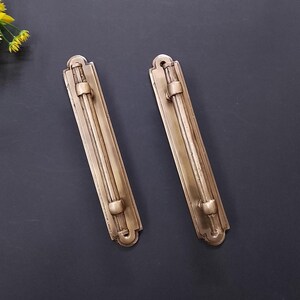 May include: Two elongated, rectangular brass cabinet pulls with a metallic finish. Each handle features a central rod mounted on a flat, decorative backplate with rounded ends and two circular supports securing the rod to the plate.
