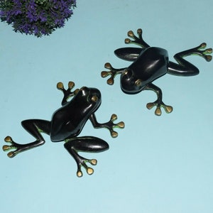 Könnte beinhalten: Zwei schwarze Froschfiguren mit goldenen Akzenten. Die Frösche sind mit ausgestreckten Beinen posiert, als würden sie springen.