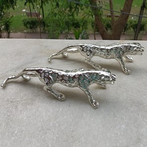 Könnte beinhalten: Zwei silberfarbene Leopardenfiguren aus Metall in schleichender Haltung. Jede Skulptur weist eine strukturierte Oberfläche mit erhabenen kreisförmigen Flecken am Körper, einen langen Schwanz und einen nach vorne gerichteten Kopf mit offenem Maul auf.