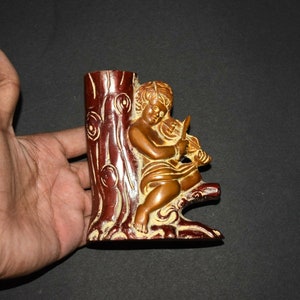 May include: A wooden figurine of a cherub playing a musical instrument, sitting on a tree trunk. The figurine is painted in shades of gold and brown.