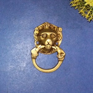May include: A brass lion head door knocker with a ring handle. The lion's face is detailed with a mane, eyes, nose, and mouth. The ring handle is held by two lion paws. The background is a solid blue.
