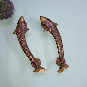 Puede incluir: Un par de tiradores de puerta decorativos con forma de pez. Los tiradores son de metal y tienen un acabado marrón. Los peces tienen escamas y aletas.