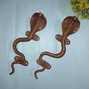 Puede incluir: Dos tiradores de puerta de latón adornados con forma de cobras con tallas detalladas. Los tiradores son de color marrón oscuro y tienen un diseño tradicional indio.