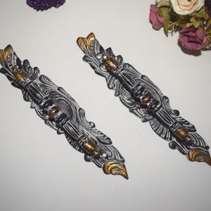 Puede incluir: Dos tiradores de puerta ornamentados en negro y dorado con intrincados diseños florales. Los tiradores son de metal y tienen un acabado desgastado.