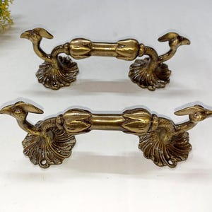 May include: Two antique brass drawer pulls. Each pull features a central bar with decorative bird-like figures at each end, mounted on ornate, scalloped bases. The brass has a warm, aged patina, suggesting vintage or antique origins.