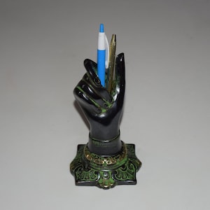 Könnte beinhalten: Eine schwarze Handskulptur mit grünen und goldenen Akzenten hält zwei Stifte, einen blauen und einen goldenen, in einem dekorativen Stifthalter.