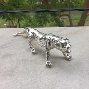 Könnte beinhalten: Eine silberne Metallfigur eines Leoparden mit gepunktetem Muster. Die Figur ist in einer Gehposition mit dem Kopf zur Seite gedreht.