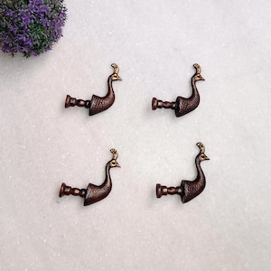 May include: Four decorative peacock-shaped drawer pulls. The pulls are a dark bronze color with gold accents on the head. The pulls are arranged on a white surface with a purple floral arrangement in the upper left corner.