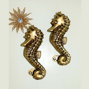 Könnte beinhalten: Zwei goldfarbene, metallene Türgriffe in Form von Seepferdchen. Die Griffe sind mit einem strukturierten, schuppigen Design verziert und haben eine kleine, ovale Basis.