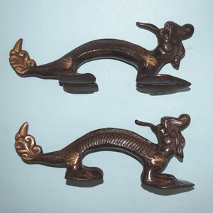 May include: A pair of antique bronze dragon door handles with gold accents. The handles are shaped like dragons with their heads turned to the side and their tails curled up. The handles are in good condition and have a beautiful patina.