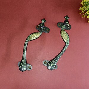 Może przedstawiać: Dwa czarne metalowe uchwyty do drzwi z zielonymi akcentami. Uchwyty mają kształt pawia z głową i ogonem skierowanymi do góry.