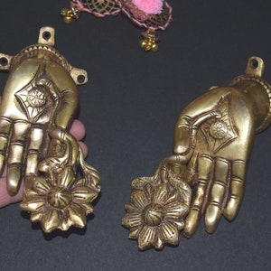 May include: Two gold-toned metal hand sculptures with a flower design. Each hand is holding a stylized flower with a circular center. The hands are detailed with lines and patterns.