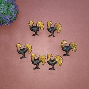 Puede incluir: Seis figuritas de gallo negras y doradas con un acabado dorado en las plumas de la cola y la cresta. Los gallos están de pie sobre una superficie roja.