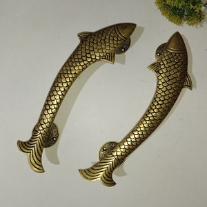 Pode incluir: Duas maçanetas de porta em forma de peixe douradas com detalhes intrincados. As maçanetas são feitas de metal e têm um acabamento polido.